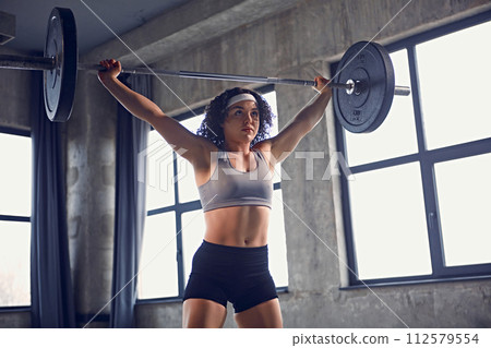 Dynamic portrait of young fit woman in sportswear practicing squat technique with barbell overhead in motion at gym. 112579554