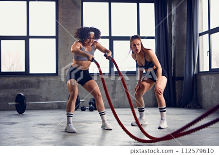 Athletic female model in fitness session with battle ropes at gym. Two fit girls wearing sportswear exercising in motion. Athletic female model in fitness session with battle ropes at gym. Two fit girls wearing sportswear exercising in motion. 112579610