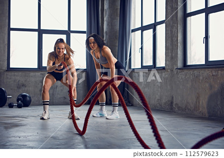 Focused female athletes, bodybuilders using heavy ropes for training in motion in industrial style fitness studio. Focused female athletes, bodybuilders using heavy ropes for training in motion in industrial style fitness studio. 112579623