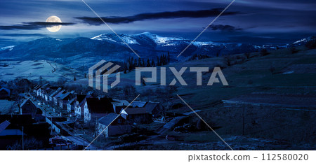 alpine countryside ant night. village in the valley and distant mountain range with snow capped tops in full moon light 112580020