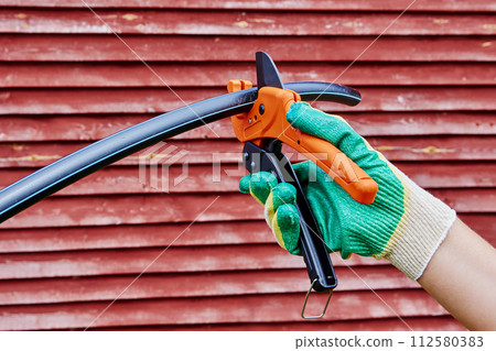 Flexible HDPE pipe is cut by plumber when installing an external plumbing system for drinking water and irrigation, close-up against background of red wooden wall. Flexible HDPE pipe is cut by plumber when installing an external plumbing system for drinking water and irrigation, close-up against background of red wooden wall. 112580383