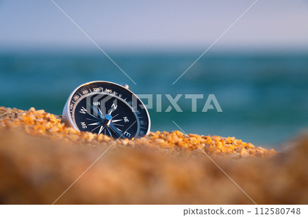 Conceptual photo of a compass in coarse sand against the background of the sea and sky, close-up, without people 112580748