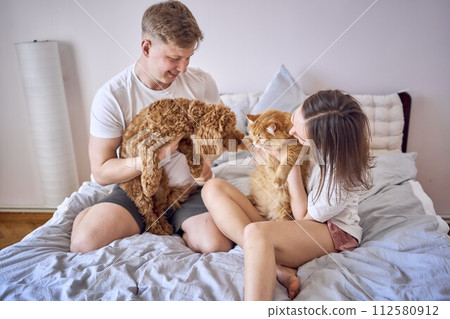 young couple spending time with their animals, a red cat and a cockapoo girl, playing and laughing young couple spending time with their animals, a red cat and a cockapoo girl, playing and laughing 112580912