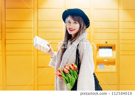 Portrait of smiling fashion woman with box near modern postal automatic mail terminal with self service device for pickup or refund an order. Electronic locker for storing parcels. Selective focus. 112584019