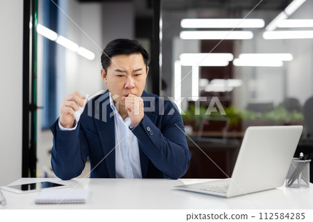 Young Asian male businessman sitting at desk in office and suffering from severe cough and asthma attack, holding napkin and covering mouth with hand. Young Asian male businessman sitting at desk in office and suffering from severe cough and asthma attack, holding napkin and covering mouth with hand. 112584285