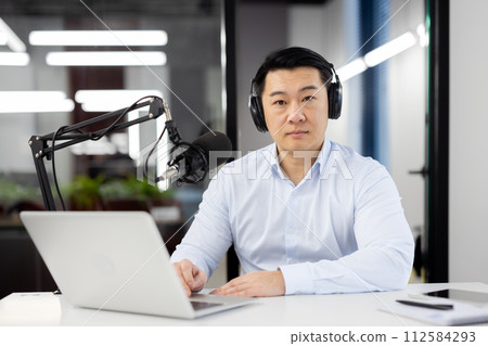 Portrait of a serious young businessman sitting in the office at the desk wearing headphones and in front of a laptop, confidently looking at the camera. 112584293