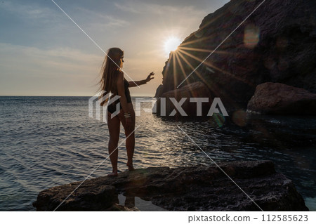 Woman tourist enjoying the sunset over the sea mountain landscape. Sits outdoors on a rock above the sea. She is wearing jeans and a blue hoodie. Healthy lifestyle, harmony and meditation 112585623