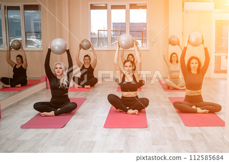 A group of six athletic women doing pilates or yoga on pink mats in front of a window in a beige loft studio interior. Teamwork, good mood and healthy lifestyle concept. A group of six athletic women doing pilates or yoga on pink mats in front of a window in a beige loft studio interior. Teamwork, good mood and healthy lifestyle concept. 112585684