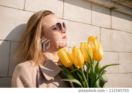 Woman holding yellow tulips, leaning against stone wall. Women's holiday concept, giving flowers. Woman holding yellow tulips, leaning against stone wall. Women's holiday concept, giving flowers. 112585703