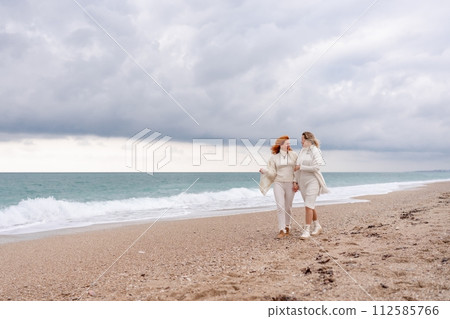 Women sea walk friendship spring. Two girlfriends, redhead and blonde, middle-aged walk along the sandy beach of the sea, dressed in white clothes. Against the backdrop of a cloudy sky and the winter 112585766