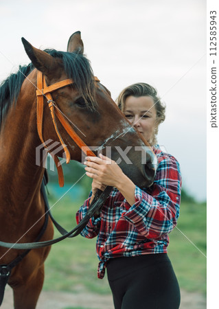 Happy blonde with horse in forest. Woman and a horse walking through the field during the day. Dressed in a plaid shirt and black leggings. 112585943