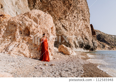 Woman red dress sea. Woman in a long red dress posing on a beach with rocks on sunny day. Girl on the nature on blue sky background. 112585948