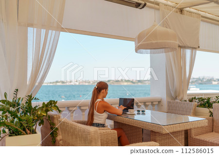 Woman coffee cafe laptop sea. Female freelancer using laptop on coffee break in modern cafe with sea view in the morning Woman coffee cafe laptop sea. Female freelancer using laptop on coffee break in modern cafe with sea view in the morning 112586015