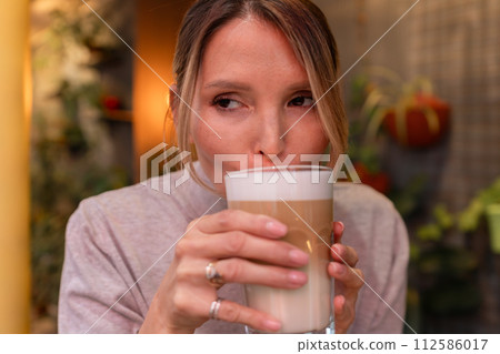 Woman with blonde hair sips cappuccino in a cafe. She is holding the glass up to her face, taking a sip of the drink. Woman with blonde hair sips cappuccino in a cafe. She is holding the glass up to her face, taking a sip of the drink. 112586017