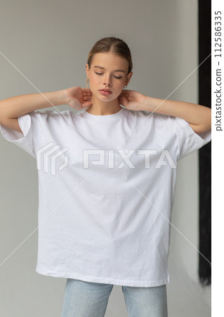 Beautiful blonde girl in a white oversized T-shirt and blue jeans posing on a gray background 112586335
