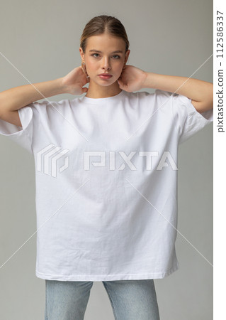Beautiful blonde girl in a white oversized T-shirt and blue jeans posing on a gray background Beautiful blonde girl in a white oversized T-shirt and blue jeans posing on a gray background 112586337