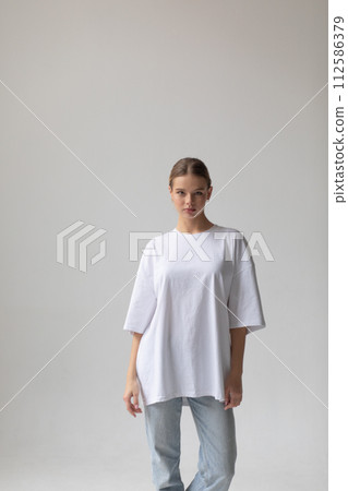 Beautiful blonde girl in a white oversized T-shirt and blue jeans posing on a gray background Beautiful blonde girl in a white oversized T-shirt and blue jeans posing on a gray background 112586379