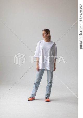 Beautiful blonde girl in a white oversized T-shirt and blue jeans posing on a gray background Beautiful blonde girl in a white oversized T-shirt and blue jeans posing on a gray background 112586392