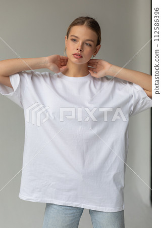 Beautiful blonde girl in a white oversized T-shirt and blue jeans posing on a gray background Beautiful blonde girl in a white oversized T-shirt and blue jeans posing on a gray background 112586396