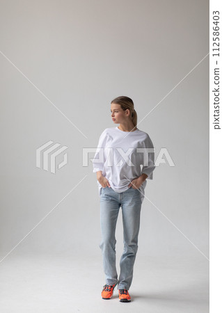 Beautiful blonde girl in a white oversized T-shirt and blue jeans posing on a gray background Beautiful blonde girl in a white oversized T-shirt and blue jeans posing on a gray background 112586403