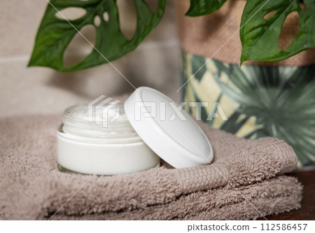 Opened cream jar with blank lid on brown towel near green monstera plant in bathroom, mockup Opened cream jar with blank lid on brown towel near green monstera plant in bathroom, mockup 112586457