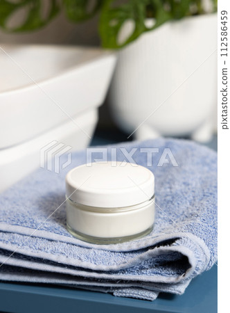 Cosmetic cream jar on blue towels near basin and green monstera in bath, lid mockup Cosmetic cream jar on blue towels near basin and green monstera in bath, lid mockup 112586459