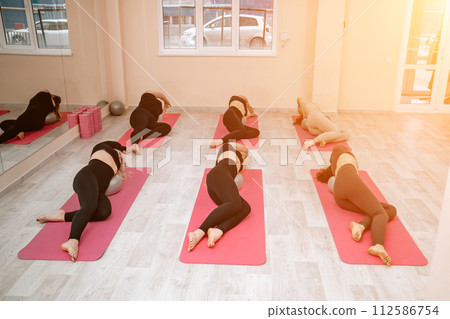A group of six athletic women doing pilates or yoga on pink mats in front of a window in a beige loft studio interior. Teamwork, good mood and healthy lifestyle concept. 112586754
