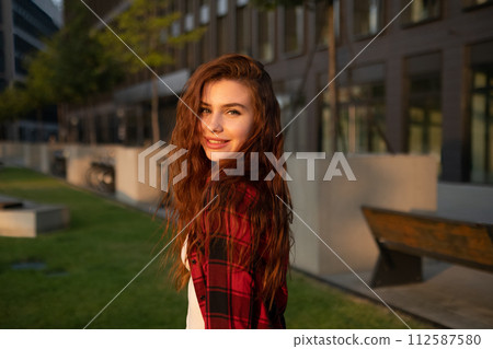 A young smiling red haired woman in a plaid shirt stands against the background of a business center 112587580