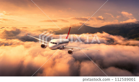 Plane is flying over mountains in low clouds at golden sunrise 112589001