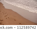 The soft glow of sunset casts a warm hue over a meandering path of footprints on the beach, alongside the gentle surf. Winding Footprints on Seashore at Sunset. 112589742