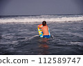 A boy in blue swim trunks turns his back to the camera, holding a vibrant inflatable float, ready to tackle the incoming wave. Boy with Inflatable Float Preparing to Ride Ocean Wave. 112589747