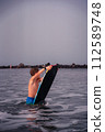 A young boy, back to the camera, walks into the calm ocean holding a bodyboard, preparing for water fun. Young Boy with Bodyboard Entering Ocean. 112589748