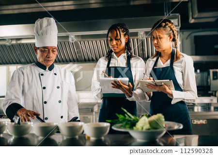 In a restaurant kitchen an Asian senior chef in uniform educates multiracial trainees. Teacher imparts cooking skills emphasizing teamwork learning and note-taking in this professional workshop. 112590328