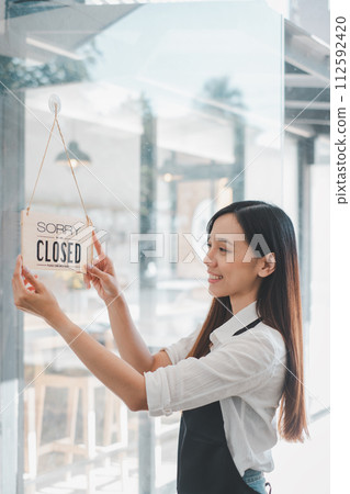 A cafe owner displays a "Sorry We Are Closed" sign with a content smile, marking the end of a fulfilling day of business in her welcoming establishment. 112592420