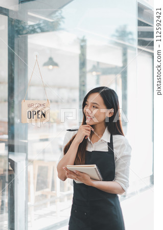 Beautiful young barista woman in apron holding order paper and standing in front of the door of cafe with open sign board. Business owner startup concept. 112592421