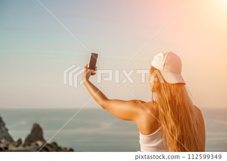 Selfie woman sea. The picture depicts a woman in a cap and tank top, taking a selfie shot with her mobile phone, showcasing her happy and carefree vacation mood against the beautiful sea background Selfie woman sea. The picture depicts a woman in a cap and tank top, taking a selfie shot with her mobile phone, showcasing her happy and carefree vacation mood against the beautiful sea background 112599349