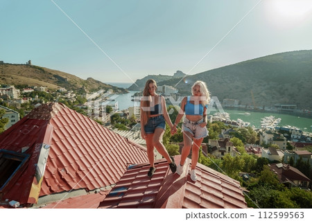 women standing on rooftop, enjoys town view and sea mountains. Peaceful rooftop relaxation. Below her, there is a town with several boats visible in the water. Rooftop vantage point. 112599563