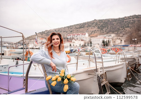 Woman holds yellow tulips in harbor with boats docked in the background., overcast day, yellow sweater, mountains 112599578