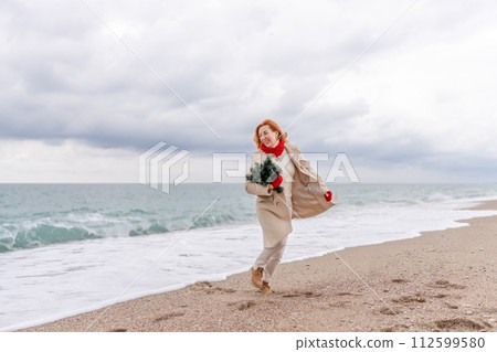 Redhead woman Christmas tree sea. Christmas portrait of a happy redhead woman walking along the beach and holding a Christmas tree in her hands. Dressed in a light coat, white suit and red mittens. 112599580
