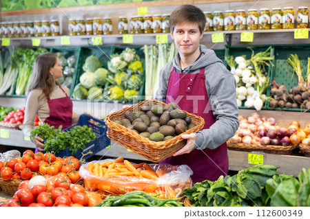 Young male seller holding avocados standing by counter in grocery market 112600349