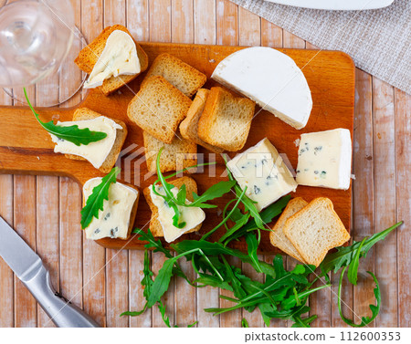 On table there is cutting board with blue cheese and hardtack decorated with arugula 112600353