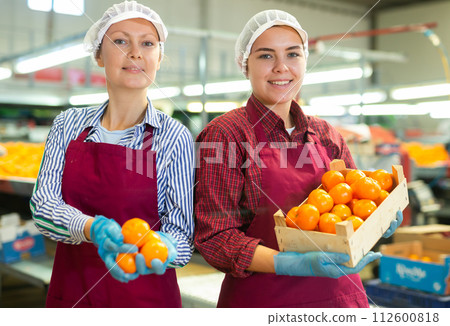 Two glad positive female employees hold a box of fresh ripe tangerines in their hands on citrus sorting line at warehouse. 112600818