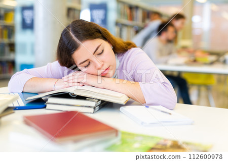 Tired young woman lying on stack of books in library 112600978