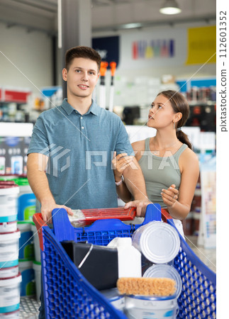 Positive married couple, young adult man and woman, shopping for materials and tools for house renovation, walking with trolley cart along aisles in hardware store 112601352