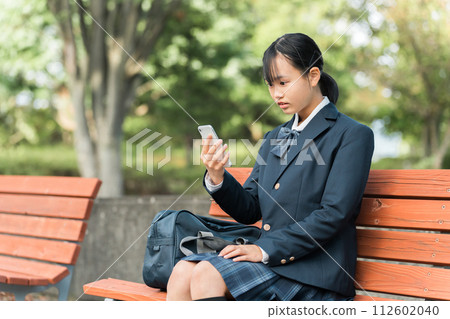 一個穿著國中或高中製服的女孩坐在公園的長椅上，在智慧型手機上觀看影片和 SNS，感到很驚訝 112602040