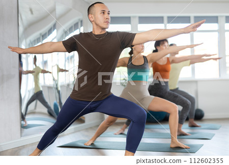 Lovers of active lifestyle on yoga mat perform doing version of warrior pose in gym Lovers of active lifestyle on yoga mat perform doing version of warrior pose in gym 112602355