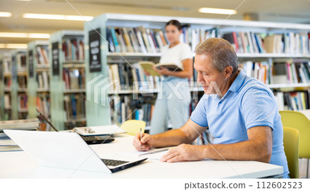 Concentrated elderly man writing in planner while searching info in books in the library Concentrated elderly man writing in planner while searching info in books in the library 112602523