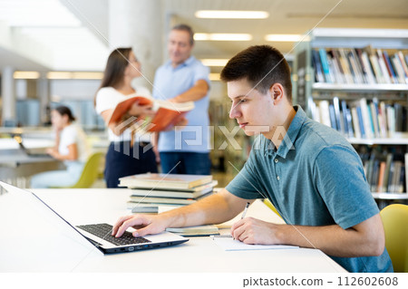 Concentrated young man checking plans in calendar while studying in the library and using laptop Concentrated young man checking plans in calendar while studying in the library and using laptop 112602608