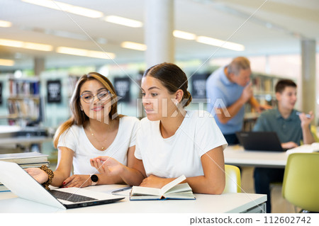 Two friendly adult female students studying in university library, using books and laptop while preparing for lecture Two friendly adult female students studying in university library, using books and laptop while preparing for lecture 112602742