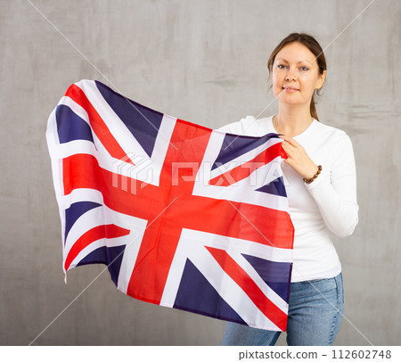 Happy young woman holding flag of Great Britain against unicoloured background Happy young woman holding flag of Great Britain against unicoloured background 112602748